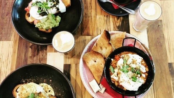 Breakfast plates from above on a timber table: eggs with avocado on toast, and shakshuka in a black pan, with lattes.