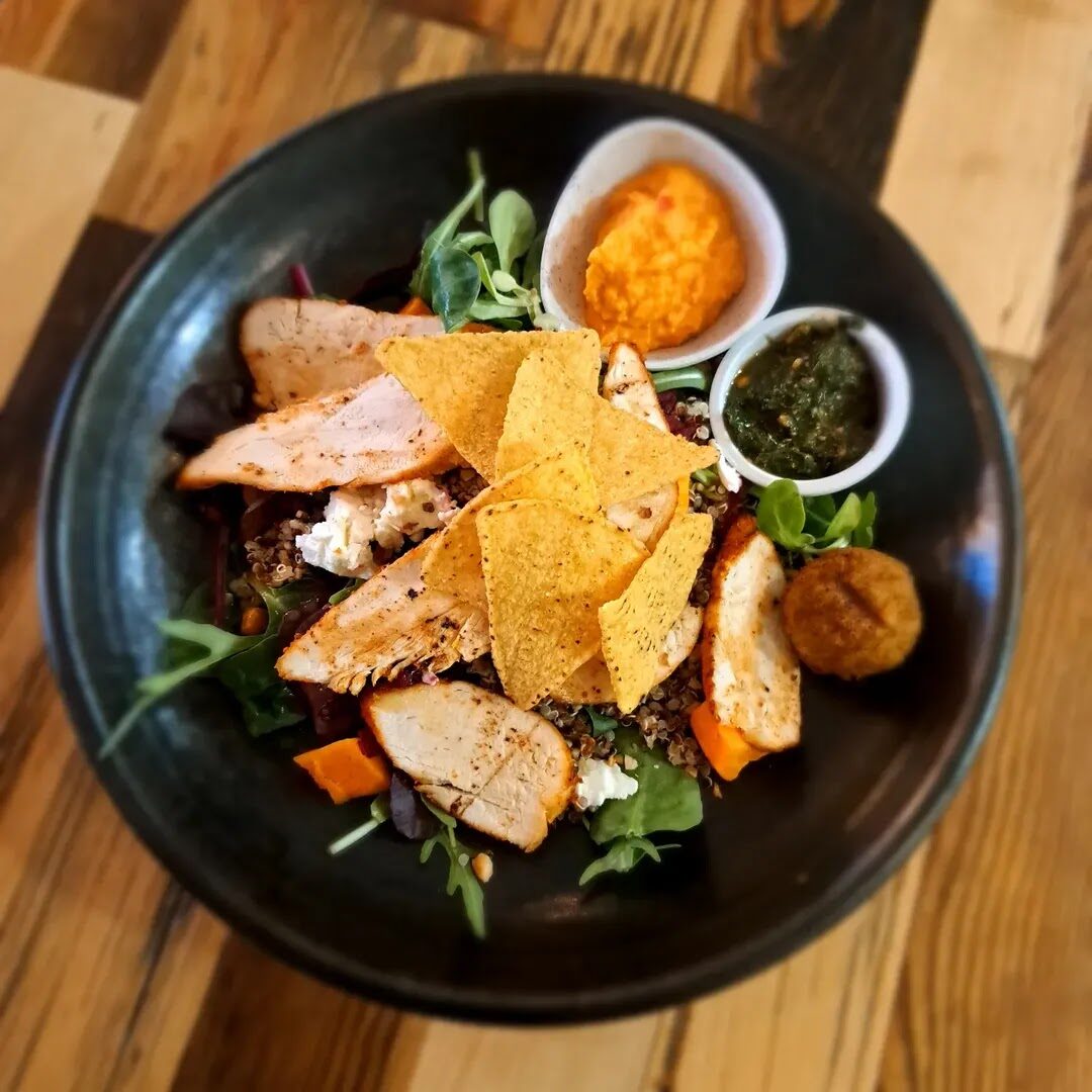 A bowl of chicken, quinoa and salad with hummus and pesto on a timber table.