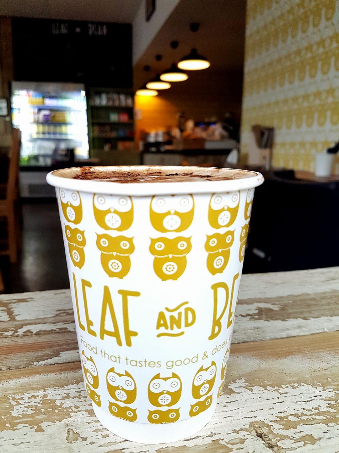 A Leaf and Bean takeaway cup printed with gold owls, on a timber table inside the café.